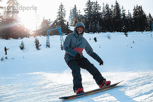 Ein Snowboarder fährt einen verschneiten Hang mit Bäumen und einem Skilift im Hintergrund hinunter.