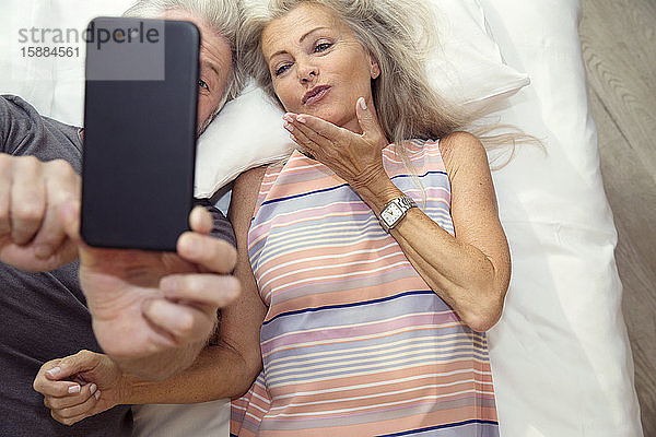 Ein Paar  das auf einem Bett liegt und sich mit einem Mobiltelefon ein Selfie macht.