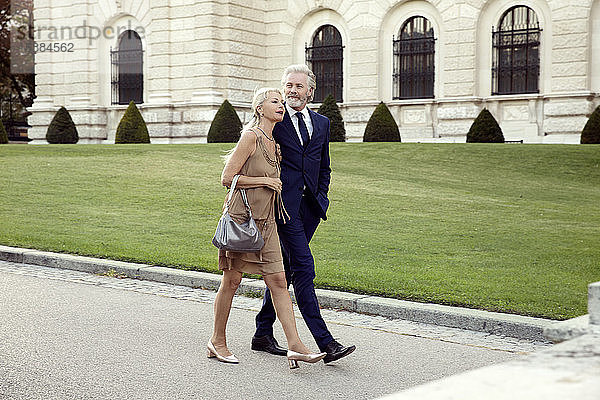 Ein Ehepaar  das Arm in Arm eine Straße entlang geht  mit einer Rasenfläche und einem großen Gebäude im Hintergrund.