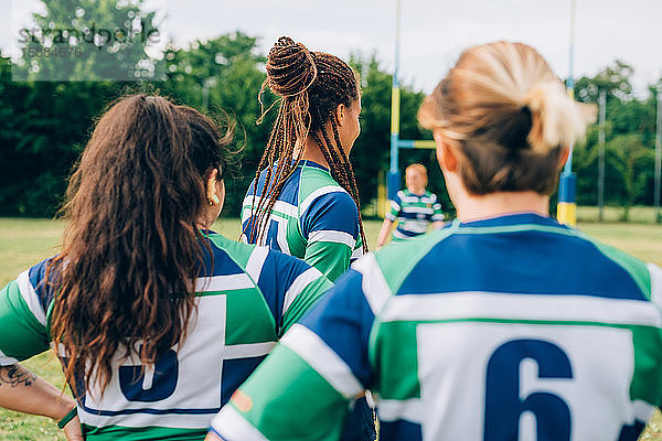 Rückansicht von drei Frauen in blauen  weißen und grünen Rugbyhemden auf einem Trainingsplatz.