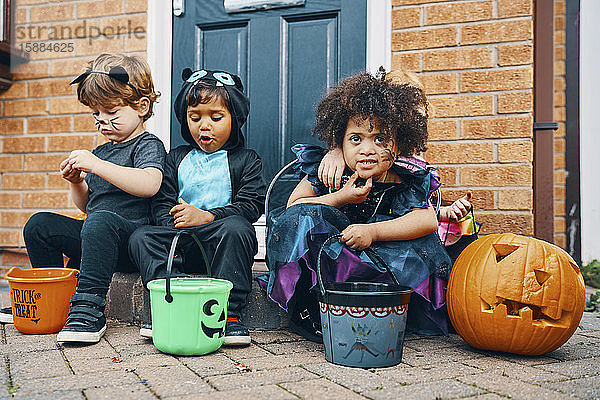 Drei für Halloween gekleidete Kinder sitzen auf einer Türschwelle und essen Süßigkeiten.
