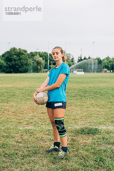 Eine Frau steht auf einem Trainingsplatz in Shorts und T-Shirt mit einem Knieband  das einen Rugbyball hält.
