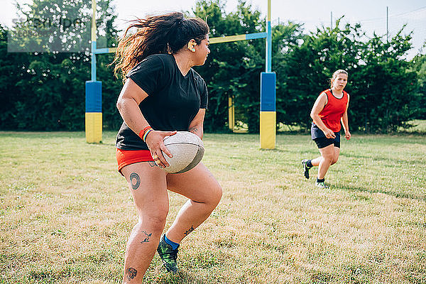 Zwei Frauen rennen auf einem Spielfeld  die eine will der anderen einen Rugbyball zuspielen.