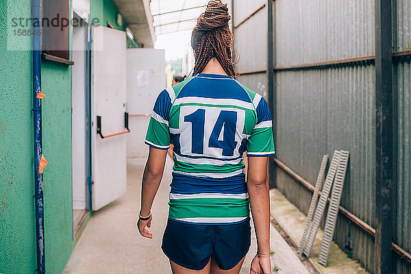 Rückansicht einer Frau in einem grün-blau-weißen Rugbyhemd  die auf die Umkleidekabinen zugeht.