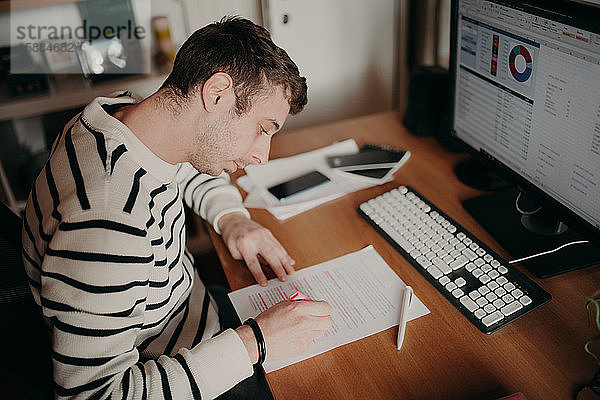 Mann  der während des Einschlusses zu Hause arbeitet und mit Stift und Papier an einem Schreibtisch sitzt.
