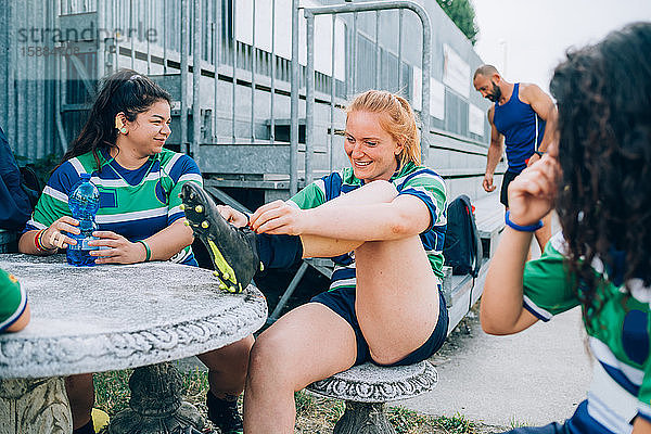 Drei Frauen in blauer  weißer und grüner Rugbykleidung sitzen an einem Tisch neben einer Metalltribüne.