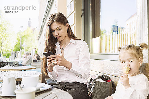 Eine Mutter und ihre Tochter sitzen vor einem Café  die Mutter am Handy  die Tochter isst ein Eis.