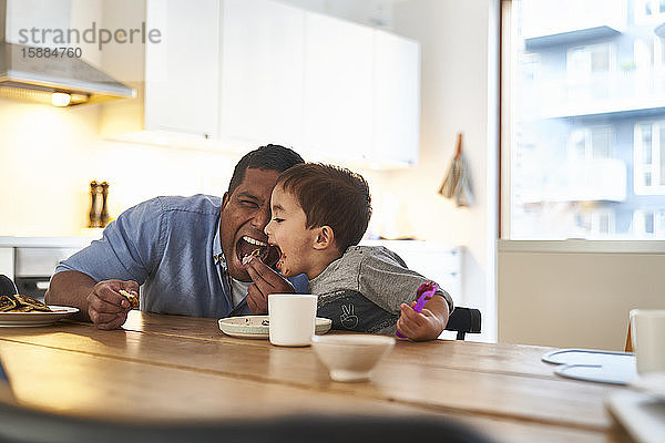 Ein Mann und ein Sohn an einem Küchentisch  beide mit weit geöffnetem Mund  essen ein Stück Schokoladenkuchen.