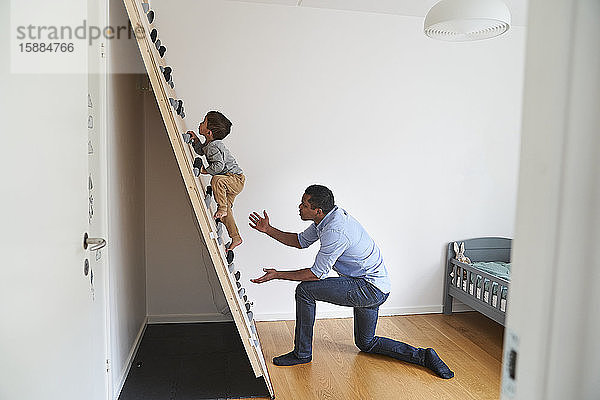 Ein Vater kniet am Boden einer Kletterwand  während sein Sohn an ihr hochklettert.