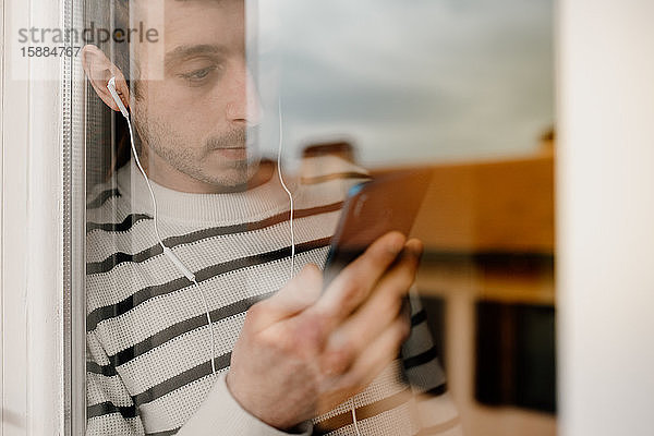 Mann  der zu Hause während der Sperrzeit arbeitet  schaut und hört mit einem Smartphone  durch ein Fenster gesehen.