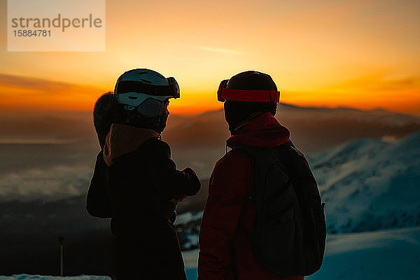 Silhouette von zwei Personen mit Skihelmen und Schutzbrille gegen Sonnenuntergang auf einem Berg im Winter
