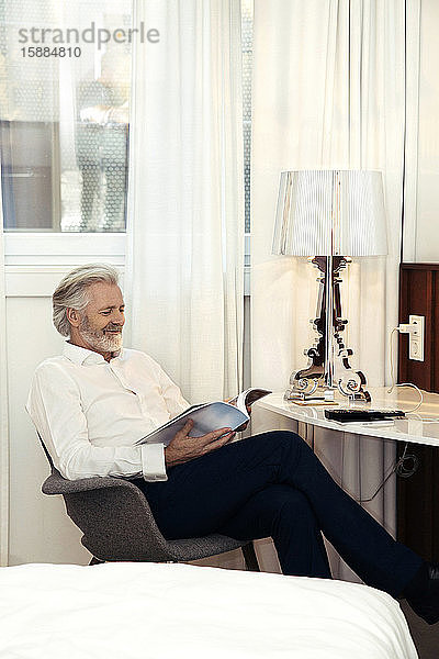Ein Mann sitzt auf einem Stuhl in einem Hotelzimmer und liest eine Zeitschrift.