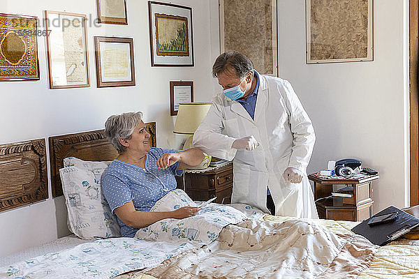 Ein Arzt im weißen Kittel und mit einer schützenden Gesichtsmaske bei einem Hausbesuch bei einer älteren Patientin  Begrüßung durch Berühren der Ellbogen.