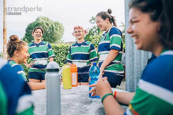 Fünf Frauen tragen blaue  weiße und grüne Rugbyhemden und lachen um einen Tisch mit 4 Wasserflaschen.