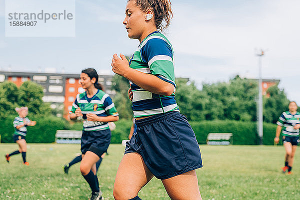 Frauen in blauen  weißen und grünen Rugbyhemden laufen auf einem Trainingsplatz.