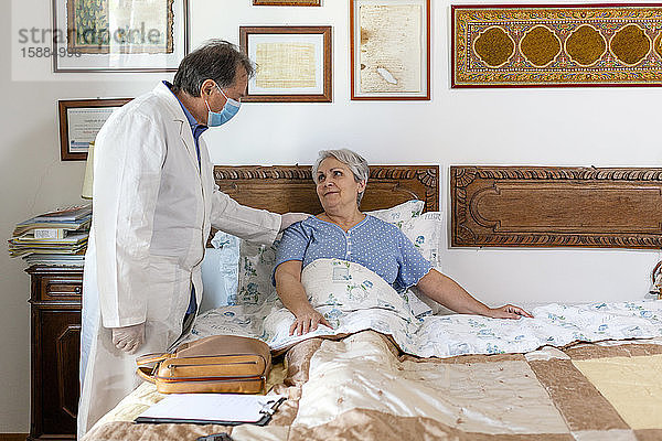 Ein Arzt im weißen Kittel und mit einer schützenden Gesichtsmaske bei einem Hausbesuch bei einer älteren Patientin.
