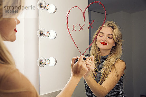 Eine Frau  die in einen Badezimmerspiegel schaut und mit Lippenstift ein Herz auf den Spiegel zeichnet.