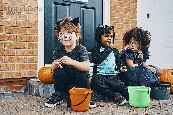 Drei für Halloween verkleidete Kinder sitzen auf einer Türschwelle und essen Süßigkeiten.