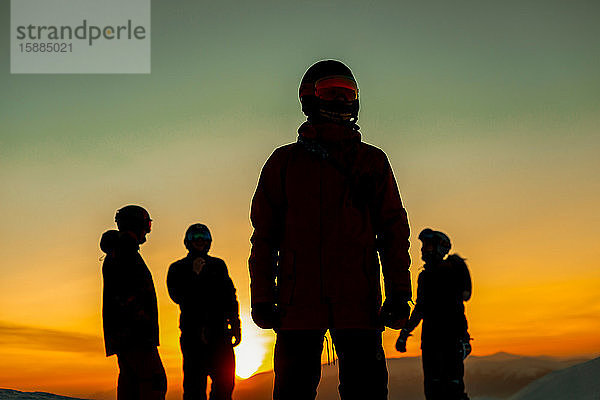 Vier Personen mit Skianzügen  Brillen und Helmen  die sich vor einem Sonnenuntergang abheben.