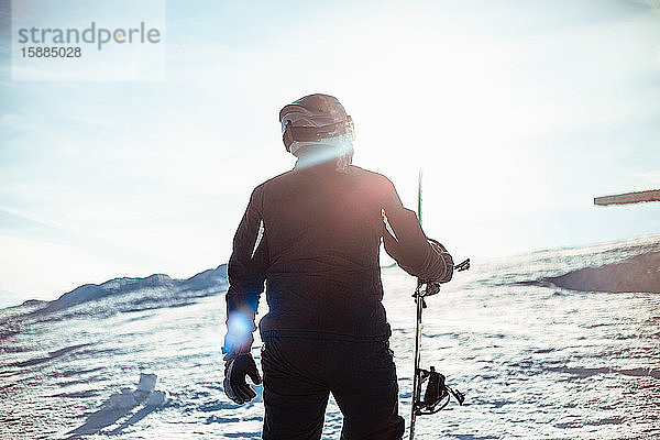 Silhouette einer Person in einem schwarzen Skianzug  mit Helm und Brille  die ein Snowboard auf einem verschneiten Hang hält.