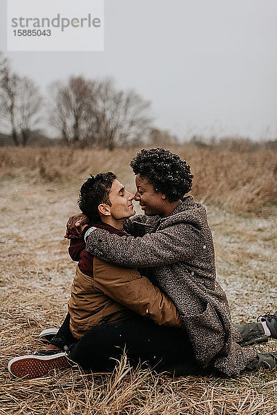 Ein weißer Mann und eine schwarze Frau sitzen in einer winterlichen ländlichen Landschaft  umarmen sich und berühren sich an der Nase.