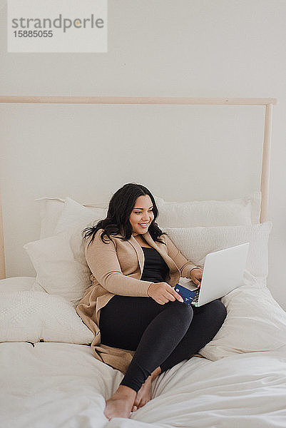 Lächelnde Frau mit langen dunklen Haaren sitzt auf dem Bett und schaut auf einen Laptop-Computer.