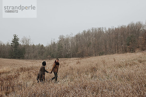 Ein Paar  das sich an den Händen hält und sich anschaut  wie es durch lange Gräser in einer winterlichen Landschaft geht.