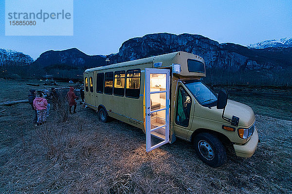 Wohnmobil in einer felsigen Landschaft in der Abenddämmerung mit Licht im Innern und offener Tür  Gruppe von Menschen steht nach hinten.