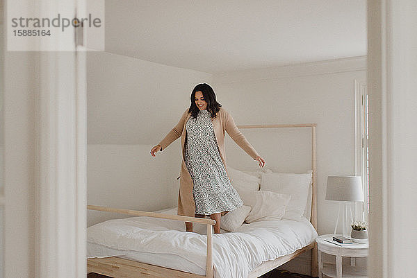 Frau mit langen dunklen Haaren springt vor Freude auf einem Bett herum.