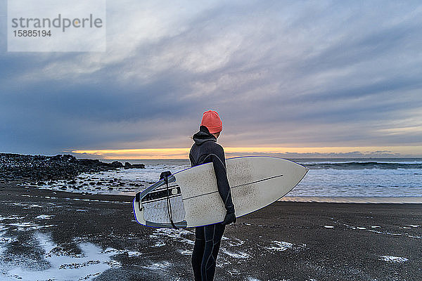 Frau in einem Neoprenanzug am Strand mit einem Surfbrett in der Hand  mit Blick aufs Meer.