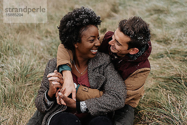Eine schwarze Frau und ein weißer Mann  die sich umarmen und lächelnd auf einem Grasfeld sitzen.