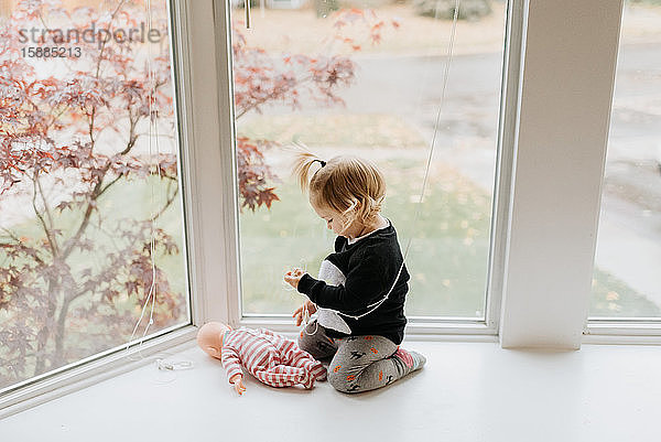 Ein kleines Mädchen in Strumpfhosen und einem Pullover  das vor einem Fenster kniet und seine Puppe vor sich auf dem Boden hat.