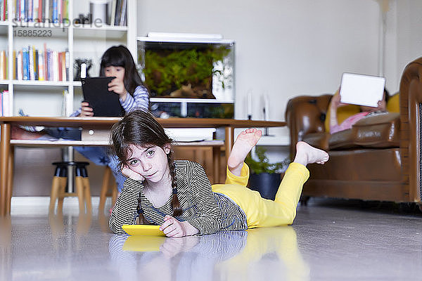 Kleines Mädchen mit Smartphone  das zu Hause auf dem Boden liegt  während ihre Schwestern im Hintergrund digitale Tablets benutzen