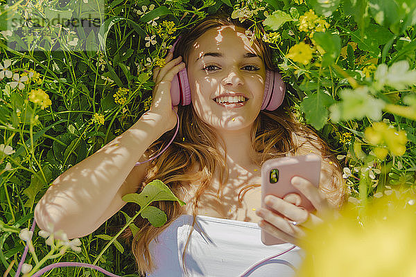 Porträt einer glücklichen jungen Frau mit Kopfhörer und Smartphone  die im Frühling auf einer Blumenwiese liegt