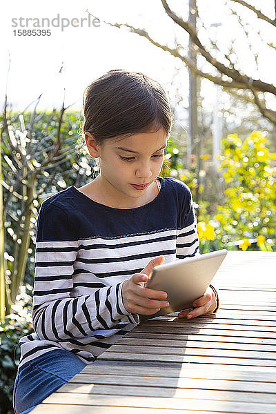 Porträt eines kleinen Mädchens  das am Gartentisch sitzt und auf ein digitales Tablett schaut