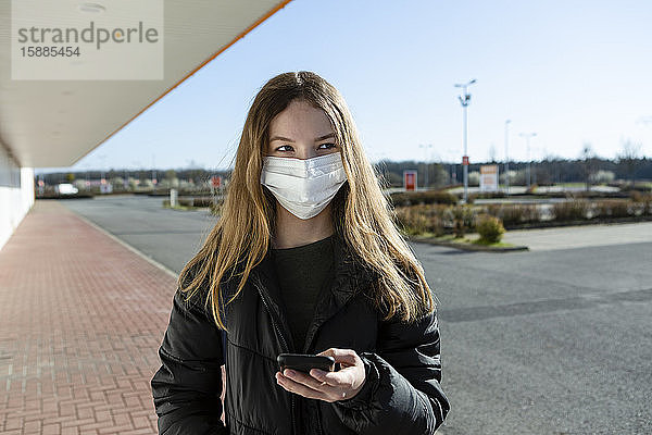 Porträt eines maskierten Mädchens in einer leeren Straße  das ein Smartphone benutzt