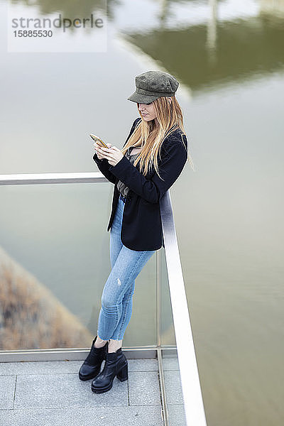 Blonde junge Frau steht auf der Terrasse vor dem Wasser und schaut auf ihr Handy