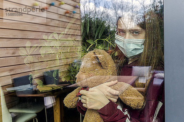 Bildnis eines Mädchens mit chirurgischer Maske und Teddybär hinter Fensterscheibe