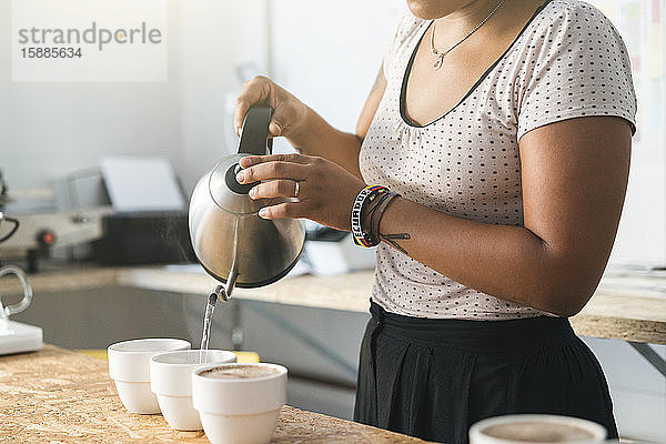 Nahaufnahme einer Frau  die in einer Kaffeerösterei arbeitet und heißes Wasser in Kaffeetassen gießt