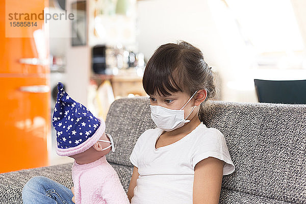 Kleines Mädchen mit Schutzmaske sitzt zu Hause auf dem Sofa und spielt mit der Puppe