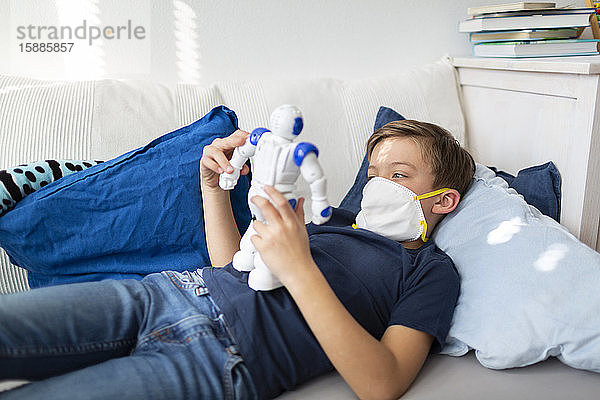 Junge mit Maske spielt während der Koronakrise mit seinem neuen  einzigen Freund  einem Roboter  in seinem Zimmer