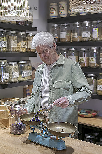 Älterer Mann hinter der Ladentheke in seinem Laden  der getrocknete Lavendelblüten in Schuppen gießt