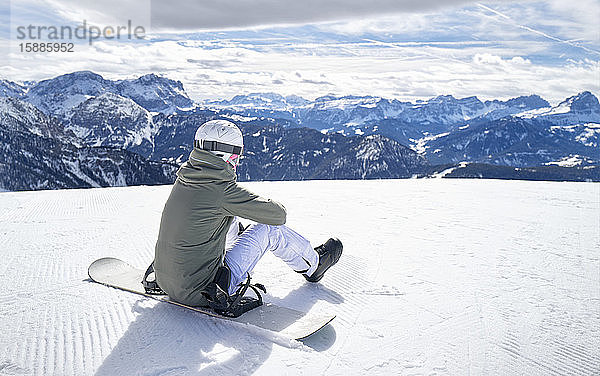 Lächelnde Frau sitzt mit dem Snowboard auf dem Aussichtspunkt und genießt die Aussicht aus der Ferne
