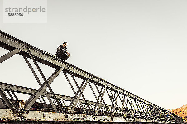 Mann kauert auf der Metallstange einer alten Eisenbahnbrücke