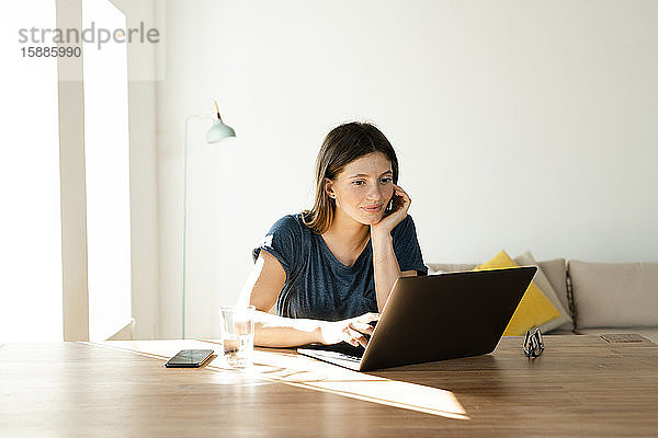 Zufriedene junge Frau arbeitet zu Hause am Laptop im Home-Office im modernen Wohnzimmer