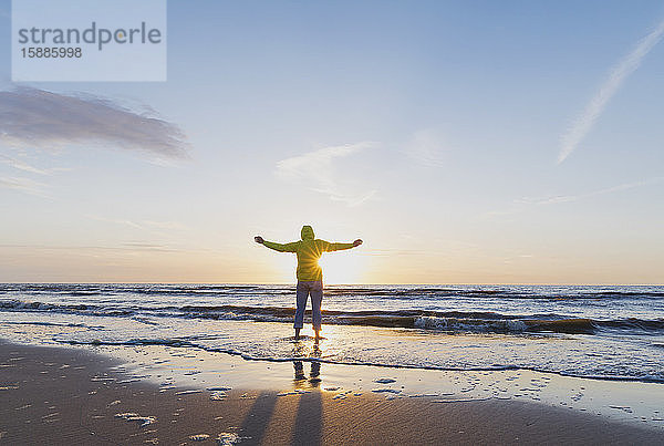 Rückansicht eines älteren Mannes in voller Länge mit ausgestreckten Armen am Ufer stehend  während er bei Sonnenuntergang das Meer gegen den Himmel betrachtet  Nordseeküste  Flandern  Belgien