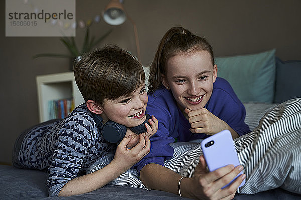 Portrait eines Bruders und seiner älteren Schwester  die zusammen auf dem Bett liegen und ein Smartphone für Video-Chat benutzen