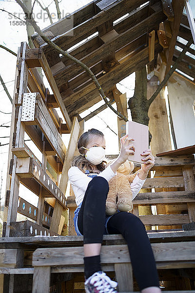 Mädchen mit Mundschutzmaske sitzt im Garten am Baumhaus  hält eine Tablette in der Hand und plaudert während der Koronaquarantäne per Videoanruf mit ihren Freunden