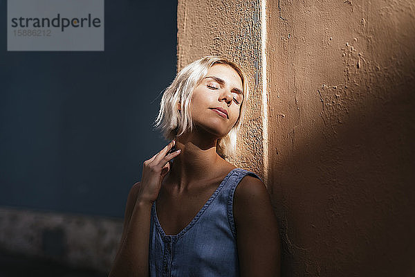 Porträt einer jungen blonden Frau  die an einer Wand lehnt und das Sonnenlicht genießt