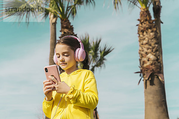 Porträt eines kleinen Mädchens in gelber Jacke  das mit Kopfhörern Musik hört und auf ein Smartphone schaut
